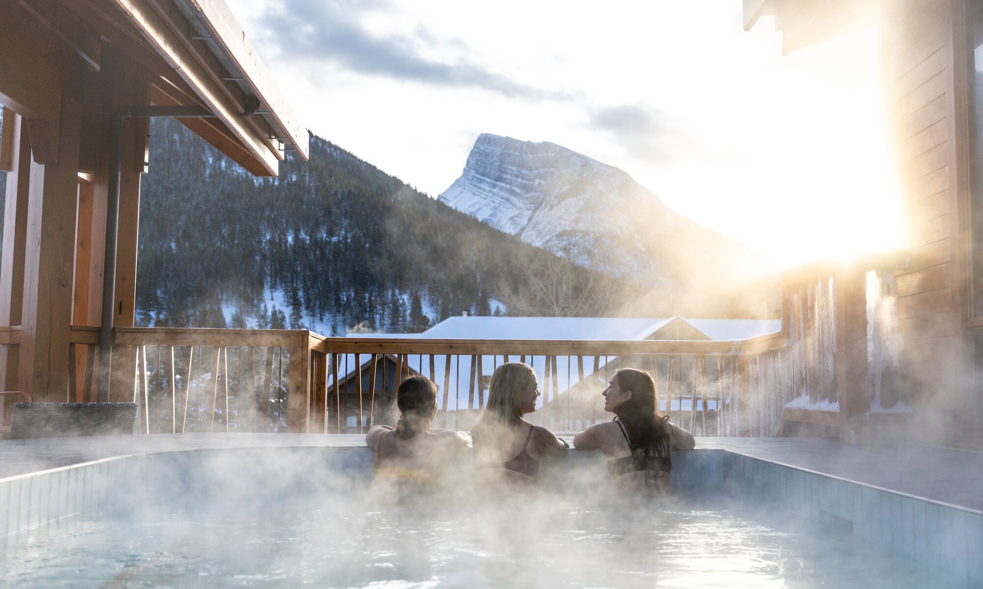 Three women hang out in the houtdoor hot tub at the Moose Hotel in Banff National Park with Mt. Rundle in the background.